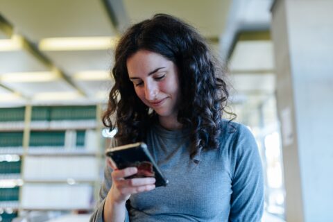 Eine junge Frau mit dunklen lockigen Haaren steht in einer Bibliothek und blickt auf ein Handy.