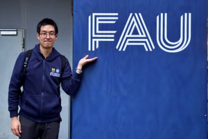 A person with a blue jacket stands next to a blue wall with the letters FAU