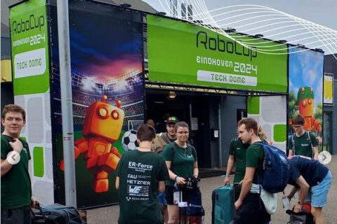 Mehrere Personen stehen vor einem großem Messestand, an dem oben ein großes grünes Banner mit der Aufschrift "Robocup Eindhoven 2024" steht. Seitlich ist ein Banner befestigt, auf dem ein Roboter mit einem Fußball gezeichnet ist.