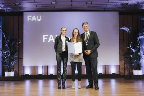 3 festlich gekleidete Personen stehen vor einem großen, blauen Bildschirm. Die in der Mitte stehende Person hat eine Urkunde in der Hand, alle lächeln freundlich in die Kamera.