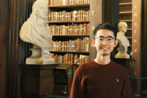 Two white busts stand in front of a shelf with books. To the side stands a person with black hair and glasses, smiling friendly into the camera.